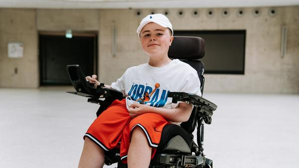 Ein Junge sitzt in einem elektrischen Rollstuhl. Er trägt Basketball-Klamotten und lächelt in die Kamera.