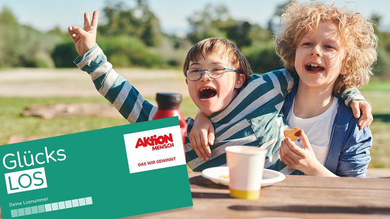 Zwei Jungen sitzen in einem Park an einem Picknicktisch und umarmen sich. Einer der beiden Jungen hat das Downsyndrom. Im Vordergrund ist ein Glücks-Los der Aktion Mensch zu sehen.