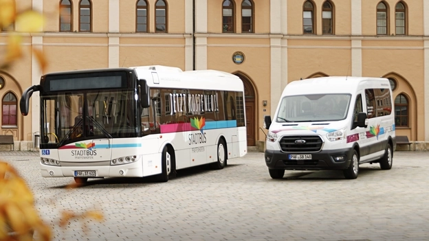Ein großer Stadtbus und ein kleinerer Sprinter-Expressbus stehen auf einem Kirchevorplatz.