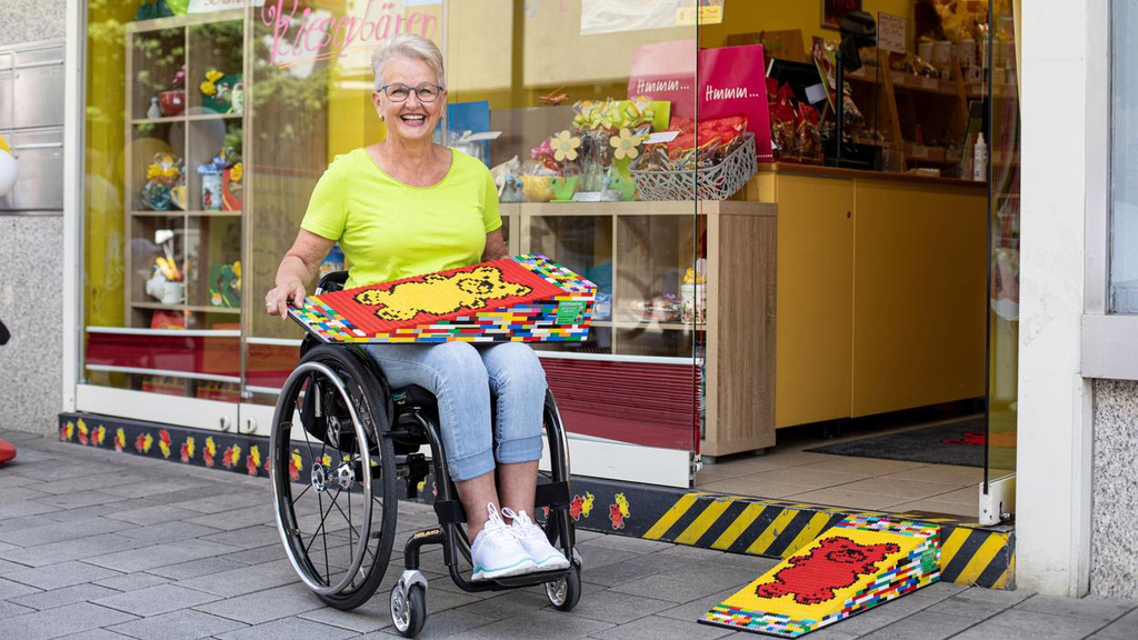 Eine Frau mit kurzen grauen Haaren und sommerlicher Kleidung sitzt in ihrem Rollstuhl vor einem Süßwarenladen und hält eine ihrer selbstgebauten Legorampen auf dem Schoß. Auf der Rampe sind bunte Gummibärchen abgebildet.