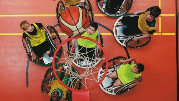 Sieben Jugendliche sitzen in Sportkleidung in Rollstühlen. Alle stehen unterhalb eines Basketballkorbs und warten auf den Treffer.