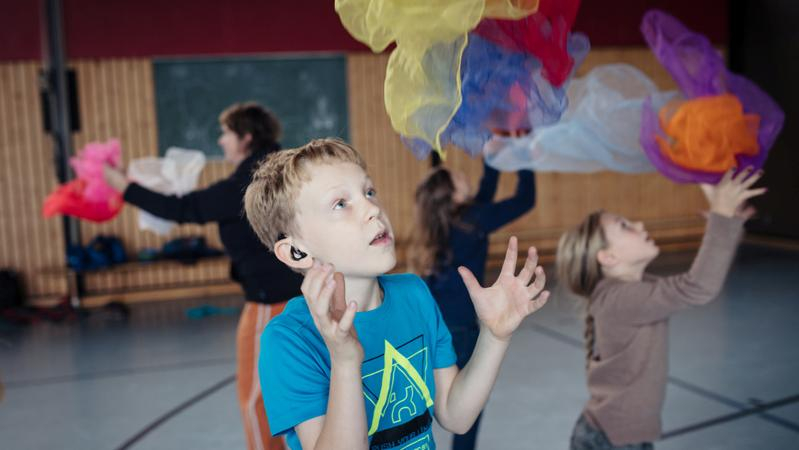 Eine Gruppe Kinder steht in einer Turnhalle und jongliert mit bunten Tüchern.