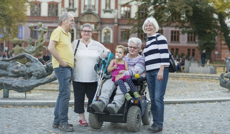 Vier Erwachsene, eine davon sitzt in einem Rollstuhl, und ein kleines Kind stehen zusammen auf einem Platz, vor einem historischen Haus, Bäume stehen im Hintergrund.
