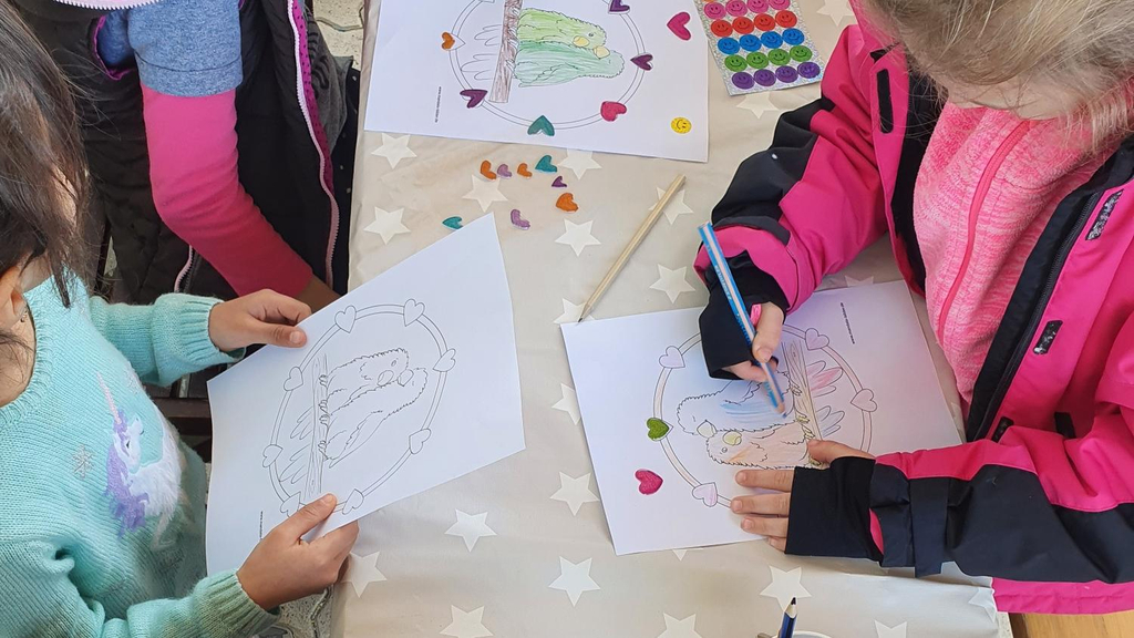 An einem Tisch sitzen Kinder, die Malbogen ausfüllen. Sie sind von oben aufgenommen und nur ihre Hände und Arme sind zu sehen.