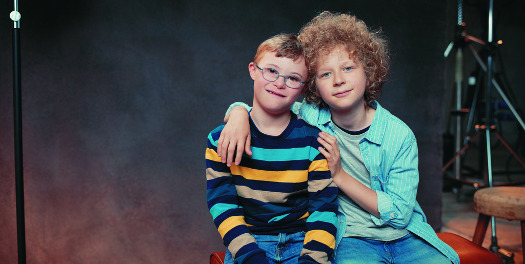 Zwei Jungen sitzen in einem Studio nebeneinander auf einer Bank. Der eine Junge legt den Arm um seinen Freund.