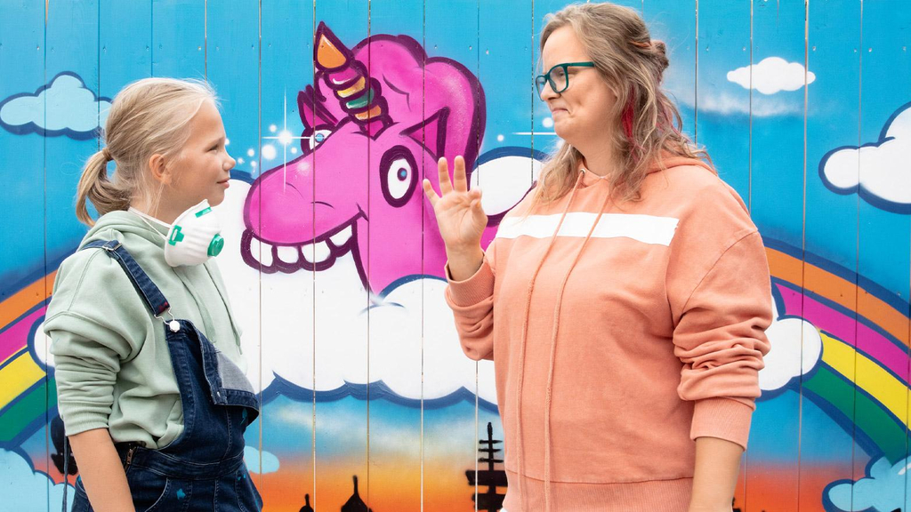 Die gehörlose Sylvia steht mit ihrer Tochter Lea vor einer Wand, auf der ein buntes Graffiti von einem Einhorn zu sehen ist.