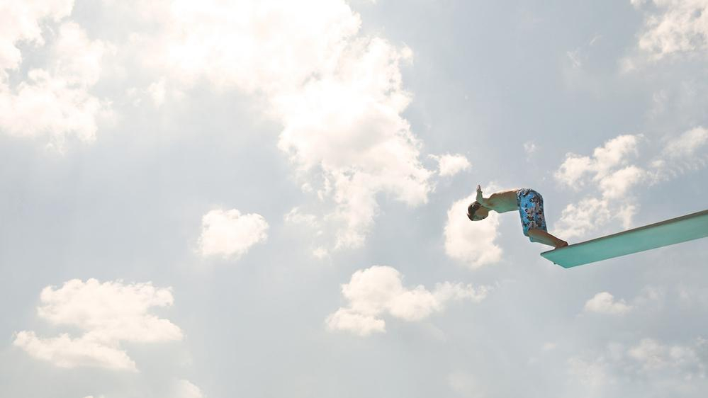 Junge in Badehose springt vom Sprungturm, Himmel und Wolken im Hintergrund