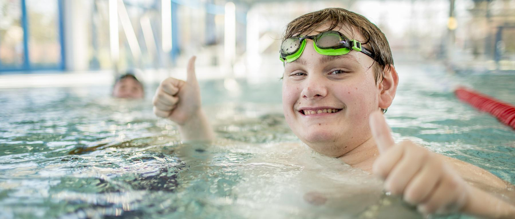 Ein Junge mit Schwimmbrille lacht aus einem Schwimmbecken heraus in die Kamera.
