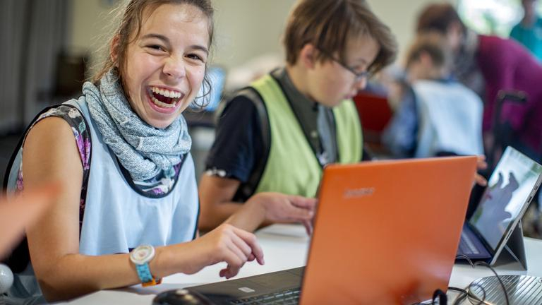 Ein Mädchen sitzt an einem Laptop und lächelt in die Kamera. Neben ihr sitzt ein Junge an einem Laptop.