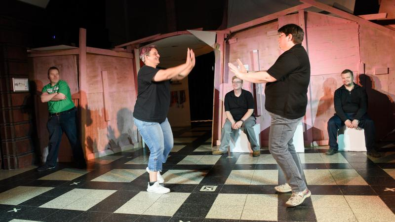 Menschen auf einer Theaterbühne, ein Paar steht mit erhobenen Händen einander gegenüber.