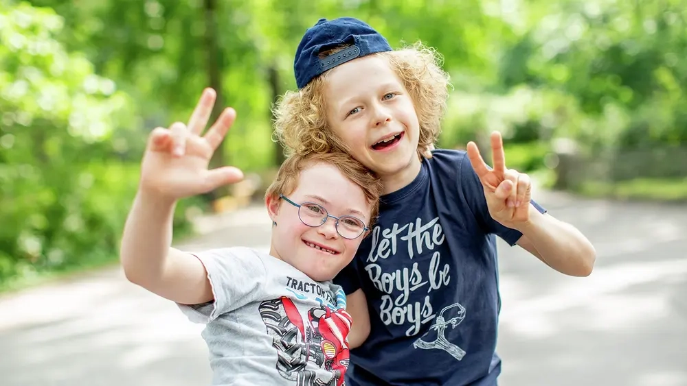 Zwei Jungen machen das Victoryzeichen und lächeln in die Kamera. Ein Junge hat das Down-Syndrom.
