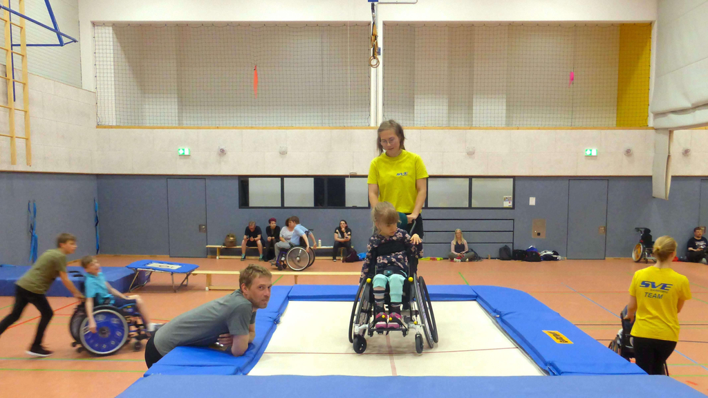 Mädchen mit Rollstuhl und Trainerin auf einem Trampolin in einer Sporthalle