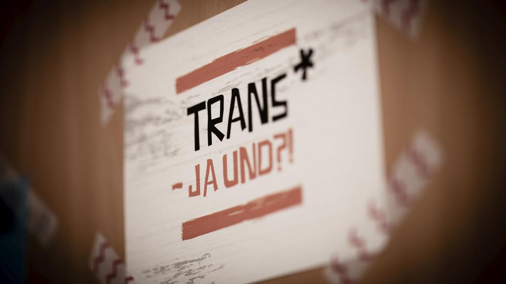Eine Postkarte klebt an einer Holztür. Darauf der Spruch: "Trans - Ja und?!".