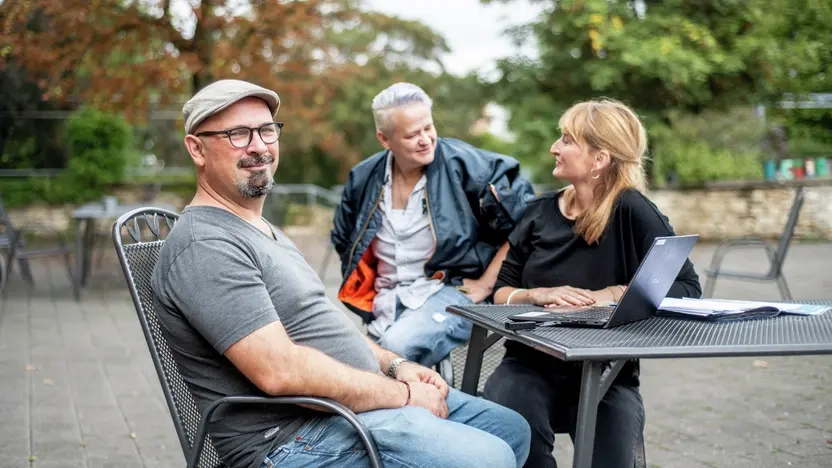 Drei Personen sitzen zusammen draußen, auf dem Tisch ist ein aufgeklappter Laptop