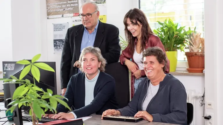 Zwei Personen sitzen vor einem Bildschirm, zwei weitere Personen dahinter stehen und schauen auch auf den Bildschirm.