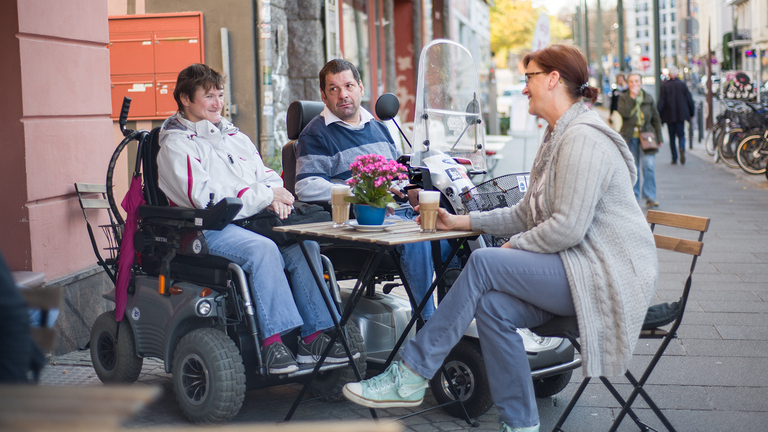Zwei Rollstuhlfahrer und eine Fußgängerin sitzen in eienm Straßencafé und trinken Kaffee