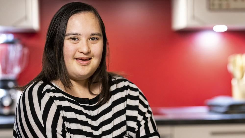 Eine junge Frau mit Down-Syndrom sitzt in der Küche ihrer WG. Sie hat die Arme verschränkt und lächelt in die Kamera.