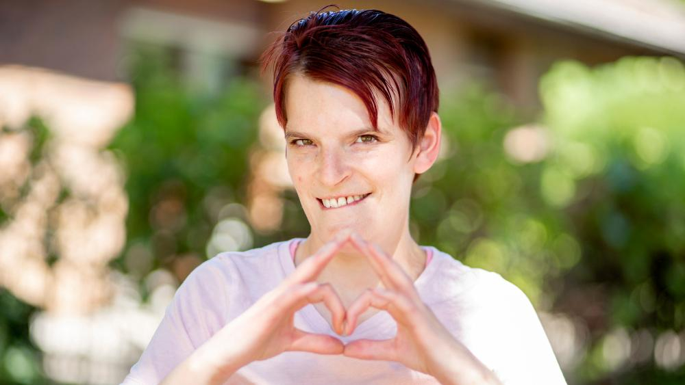 Eine junge Frau lächelt in die Kamera und bildet mit ihren Händen ein Herz.