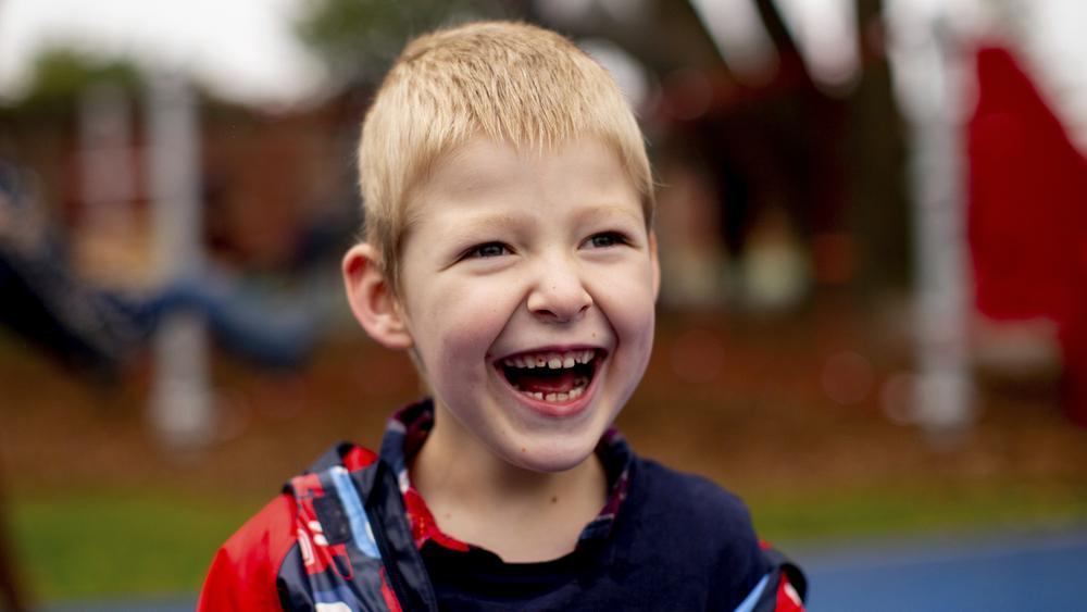 Ein Junge mit kurzem blonden Haar steht vor einem Spielplatz und lacht fröhlich.