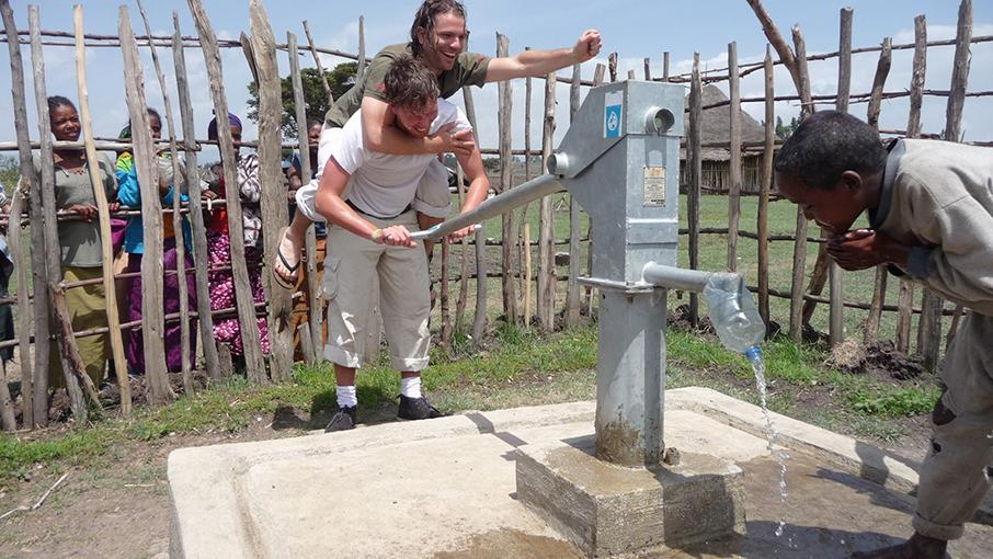 Benny huckepack auf einem Kollegen, der eine Viva con Agua Wasserpumpe betätigt. Ein Einheimischer trinkt.