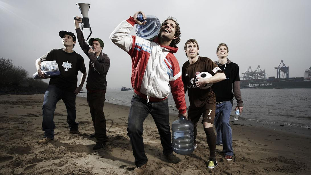 Inszeniertes Foto einer Gruppe junger Männer am Strand mit Wasserkanistern und Megafon in der Hand.