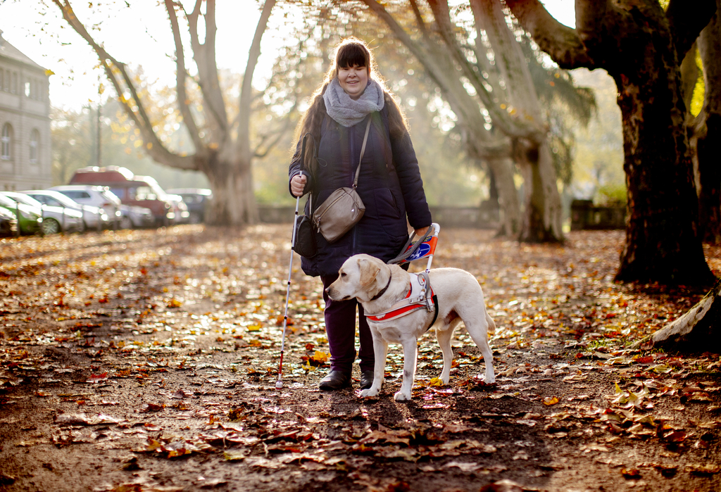 Ein Frau steht mit ihrem Blindenhund auf einem Fußweg.