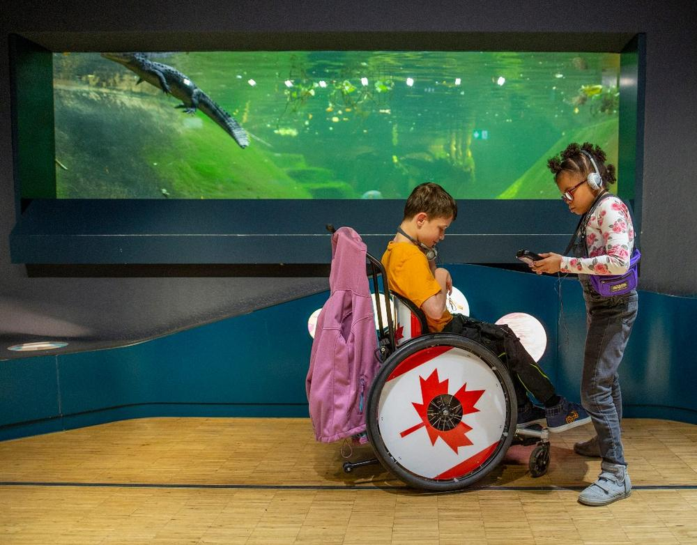 Ein Junge im Rollstuhl und ein Mädchen mit Kopfhörer stehen vor einem Aquarium.