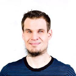 Ein junger Mann mit braunen Haaren und Bart schaut in die Kamera. Er trägt ein schwarz blau gestreiftes T-Shirt.