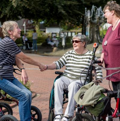 Drei Damen mit Rollstuhl und Rollator lachen und begrüßen sich auf einem Platz