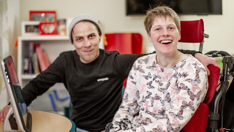 Eine junge Frau und ein junger Mann sitzen nebeneinander vor einem Computer. Die Frau sitzt im Rollstuhl.