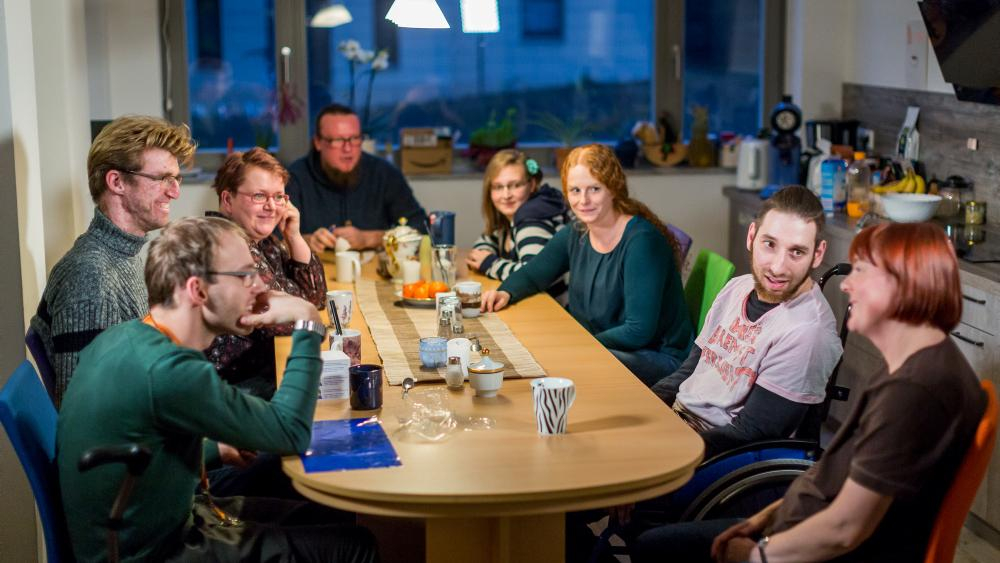 Menschen mit und ohne Behinderung sitzen an einem Tisch und lächeln in die Kamera.