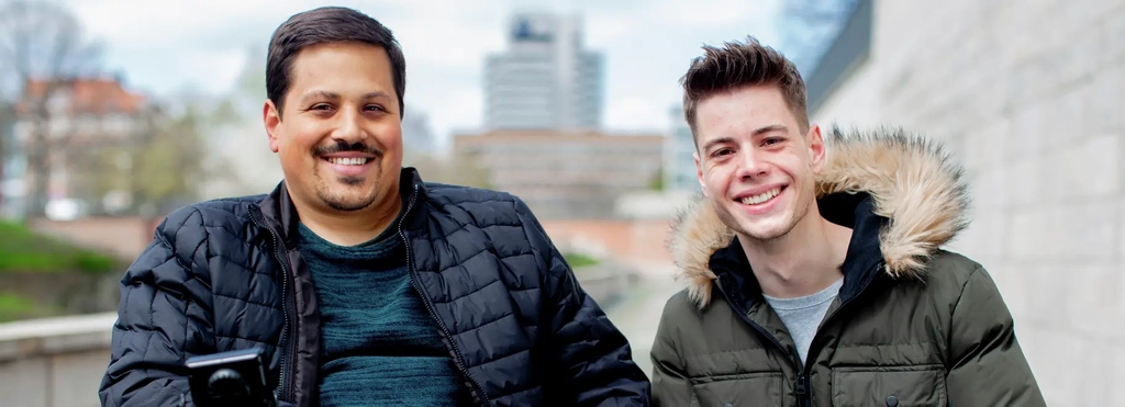 Zwei Männer sitzen lächelnd nebeneinander, einer davon im E-Rolli