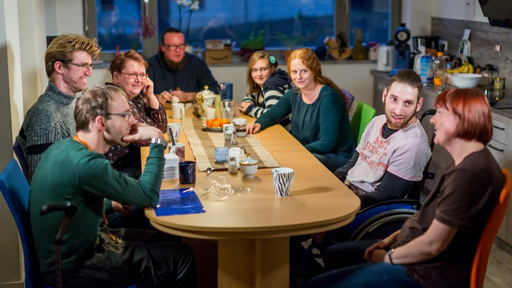 Menschen mit und ohne Behinderung sitzen an einem Tisch und lächeln in die Kamera.