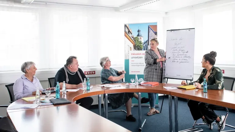 Vier Personen sitzen an einem Tisch und hören einer Referentin zu, die am Flipp-Chart etwas erklärt