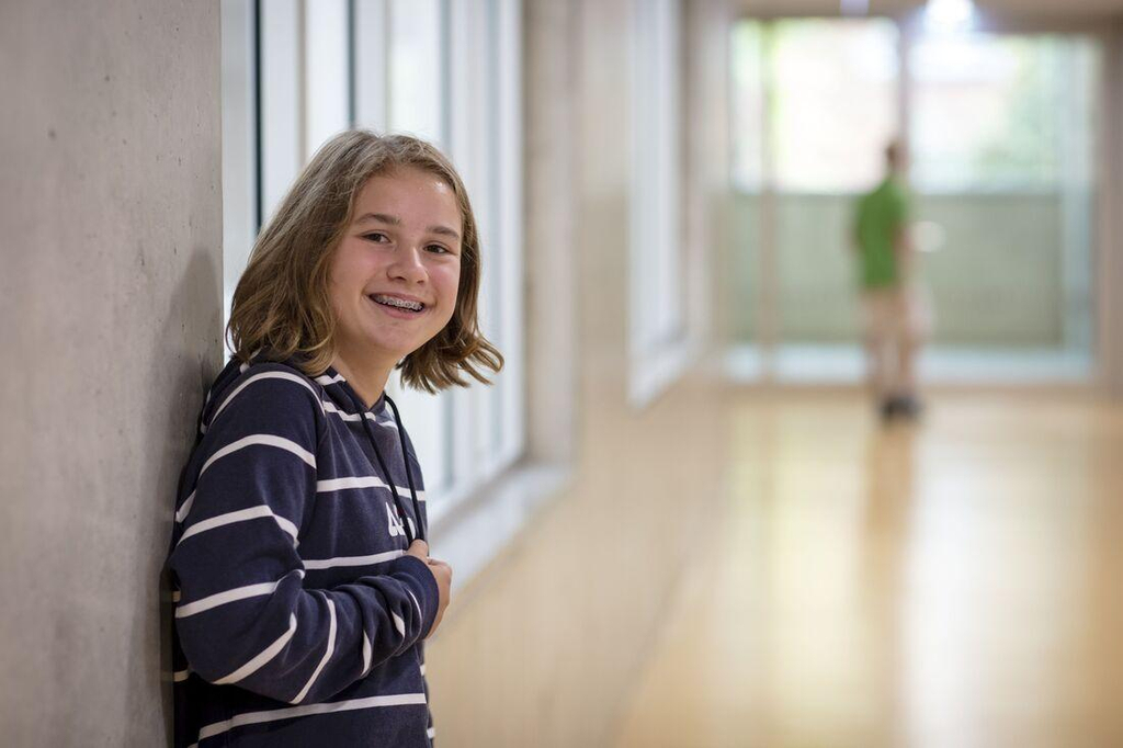 Ein Mädchen in einem gestreiften Pullover lehnt an der Wand im Flur einer Schule und lächelt in die Kamera. 