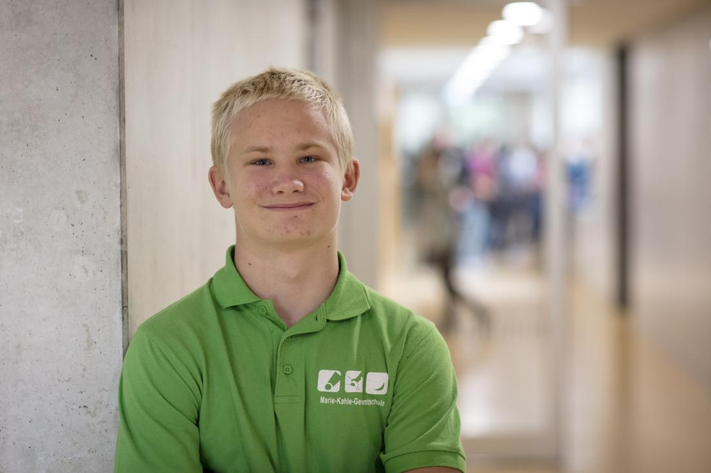 Ein blonder Junge in einem grünen Poloshirt steht im Flur eines Schulgebäudes und lächelt in die Kamera. 