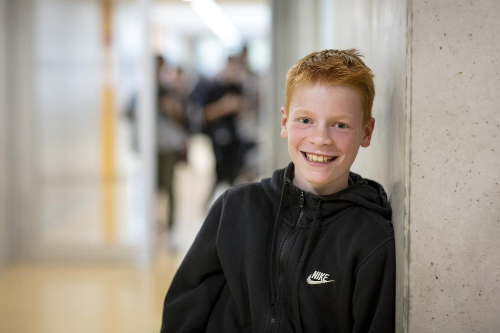 Ein rothaariger Junge in schwarzem Kapuzenpullover lehnt an einer Wand im Flur einer Schule. 