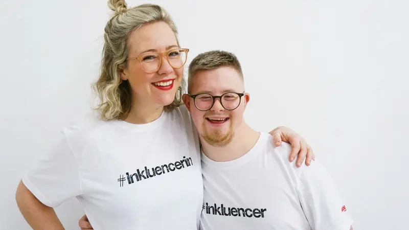 Tabea und Marian von notjustdown stehen Arm in Arm nebeneinander. Beide lachen und tragen Inkluencer-Shirts.